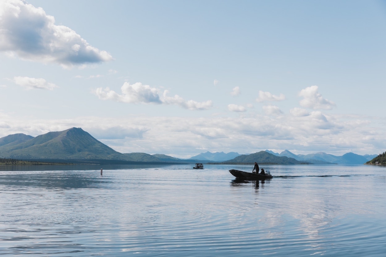 A serene lake with a clear sky, where a small boat with a person aboard glides through the calm waters, with a backdrop of distant mountains and sparse clouds.