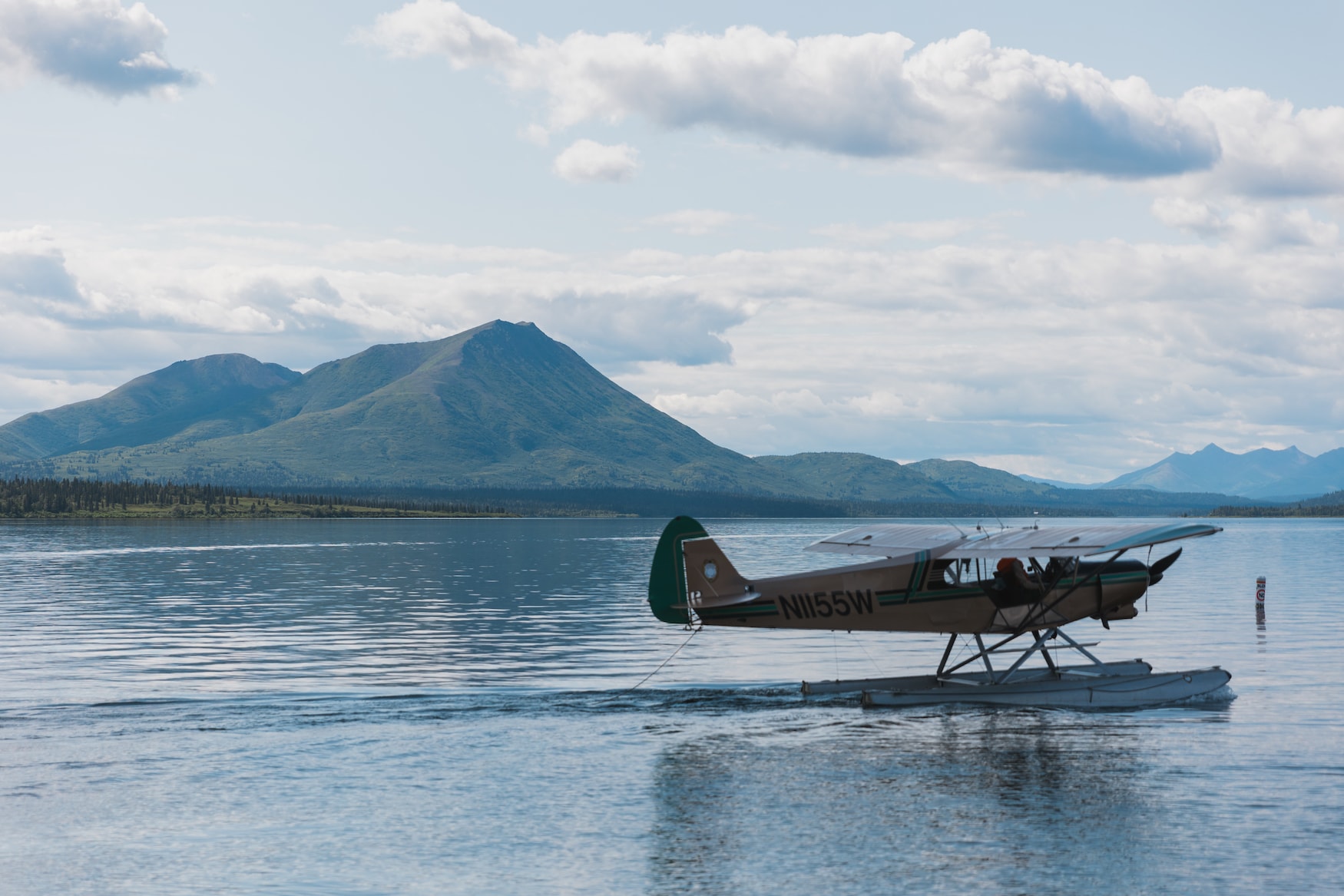 A single-engine seaplane with registration N1155W floats on a tranquil lake with a striking mountain range backdrop and scattered clouds in the sky, evoking a sense of adventure in the wilderness.