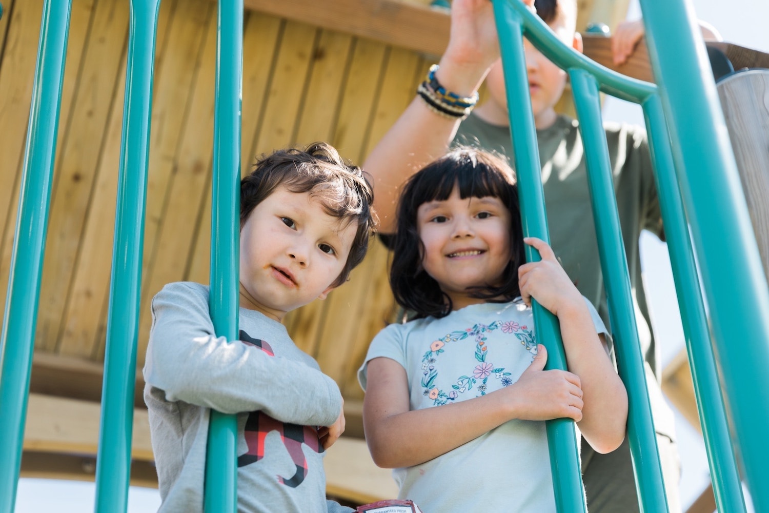 Two children playing on a playground, with a focus on a young boy and girl standing behind blue metal bars on a play structure, both looking towards the camera.