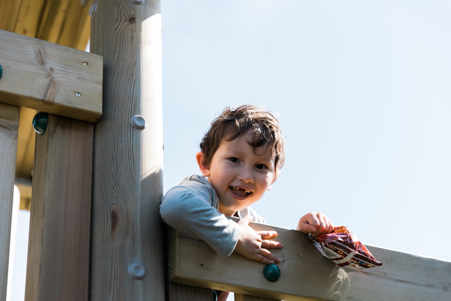 A joyful young boy leaning over the edge of a wooden play structure, with a clear blue sky in the background, holding a snack packet in his hand.
