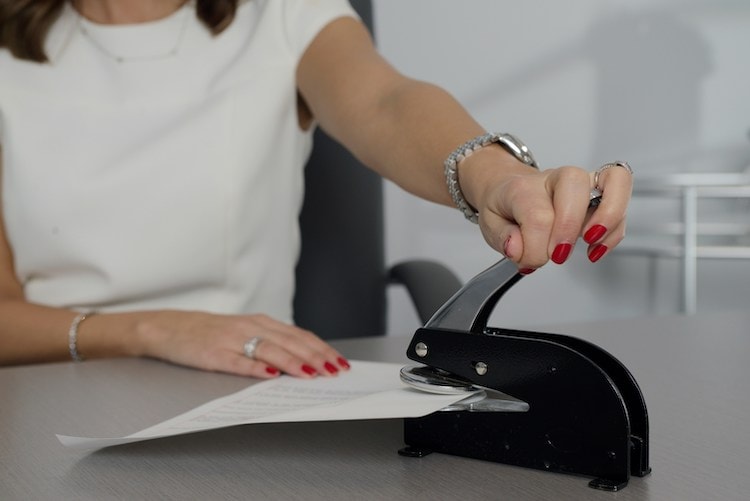 A person is using a black, heavy-duty notary public embosser on a document at a desk. Only the person's hands are visible, one holding down the paper and the other pressing the embosser, with a focus on the action of notarizing a document. Photo by Stephen Goldberg on Unsplash.